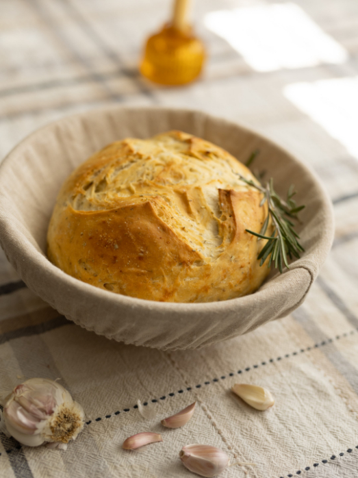 A round loaf of crusty bread in a cloth-lined basket with sprigs of rosemary. Garlic cloves are scattered on the fabric tablecloth beside it.